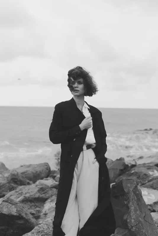 Woman wearing a long dark coat stands on a rocky shore, windswept hair, monochrome tones, ocean backdrop, stylish outdoor fashion mood with dramatic contrast