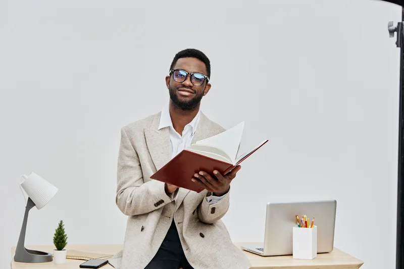 Professional young smiling man with glasses in formal beige blazer holding an open notebook while standing at a modern desk with a laptop, white background, studio shot. Business lifestyle concept