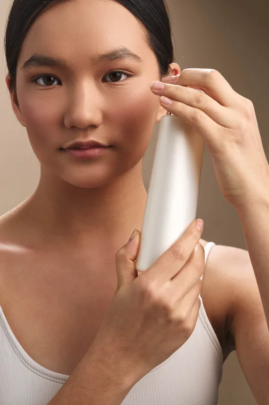asian woman gently applies skincare product from a bottle in a clean studio setting. she smiles softly, focused and calm, presenting a modern beauty routine and confident selfcare.
