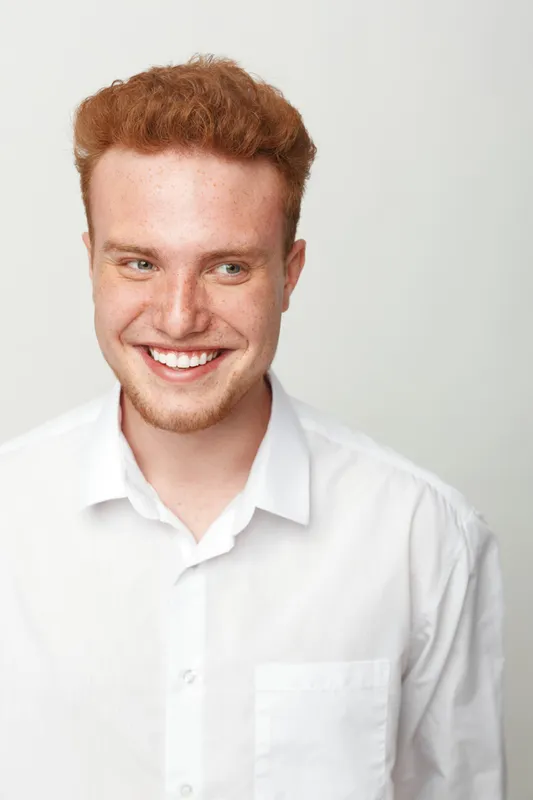Smiling young man with red hair and freckles wears white shirt, looking away with happy expression. Studio portrait on light background. Concept of youth, confidence, and natural beauty.