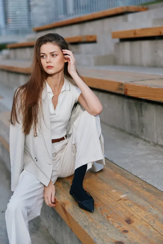 Young woman in beige suit sitting thoughtfully on wooden stairs with neutral city background, showcasing business style and calm mood KEY: woman business suit sitting thoughtful fashion urban professi