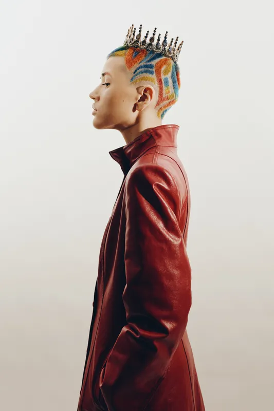 Young woman with colorful shaved hair and creative crown, wearing a stylish red leather jacket against a neutral background, showcasing bold fashion and self-expression.