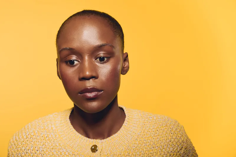 Portrait of a thoughtful black woman with dark skin wearing a yellow sweater against a yellow background. Concept of contemplation, beauty, and natural skin glow in studio lighting.