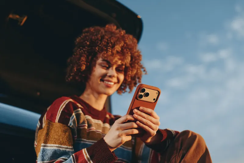 Woman lifestyle portrait outdoors, curly hair woman smiling while holding smartphone beside a car, casual striped sweater, daylight and blue sky background, relaxed mood for modern communication