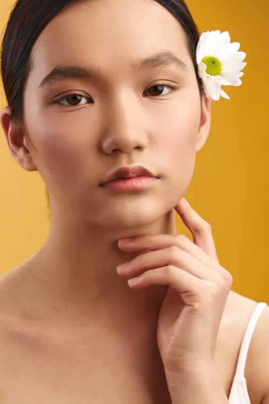 Asian woman with a white flower tucked behind her ear gazes softly at the camera against a warm yellow backdrop, presenting serene natural beauty and confident poise.