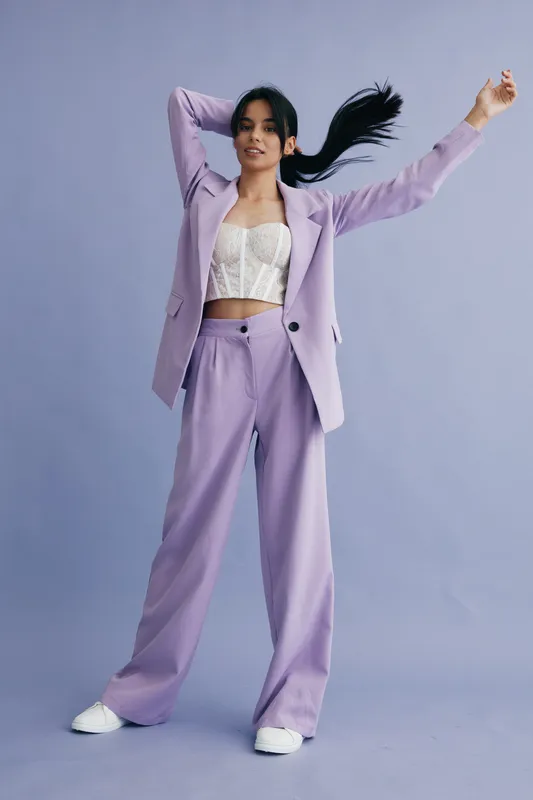 Young woman wearing a lilac suit and white crop top, posing with raised arms against a pastel purple background in a modern fashion style.
