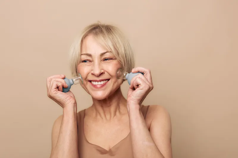 Smiling mature woman with short blonde hair uses facial massagers on cheeks, enjoying skincare routine. Beige background and natural light create warm and positive mood for beauty concept.