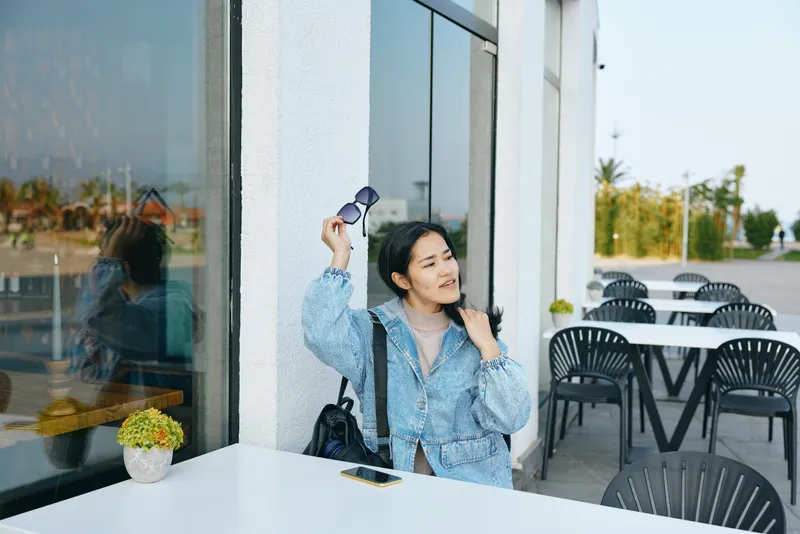 Woman, sunglasses and cafe terrace scene by glass window, stylish denim jacket, casual pose as she adjusts glasses near table, relaxed urban coffee shop setting with plants and empty chairs