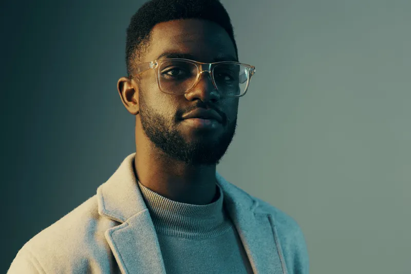 Portrait of a stylish young African man with glasses, wearing a light grey blazer over a turtleneck, showcasing confidence and modern fashion in a cool toned background