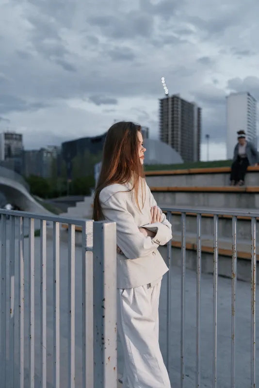 Woman in business attire standing thoughtful outdoors with city buildings and cloudy sky background in cool tones, expressing reflection and professionalism