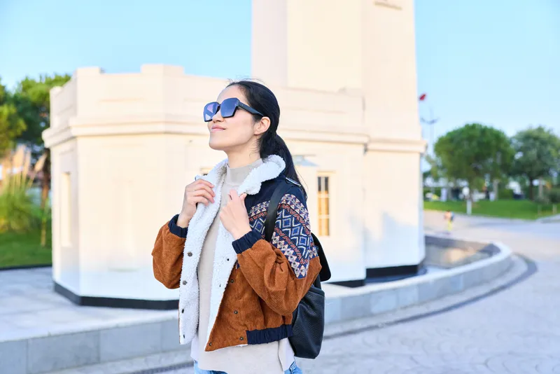 Woman wearing sunglasses, scarf and jacket, outdoors beside a monument, showcasing fashion, style and confidence during a casual stroll in daylight, urban mood, personal expression and chic accessory