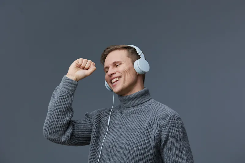Happy young man enjoying music with headphones, smiling and dancing. Casual gray sweater, positive mood, modern lifestyle, isolated on plain dark gray background. Concept: joy of listening to music.