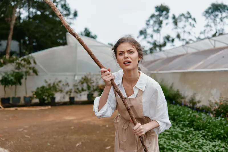 Young woman holding a stick in a garden expressing determination, wearing a casual outfit, with green plants and greenhouse in the background