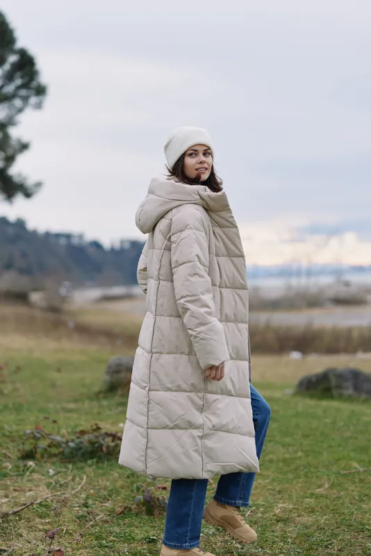 Winter woman wearing a long beige puffer coat and beanie in a grassy coastal setting, casual outdoors fashion and cozy style for chilly days.