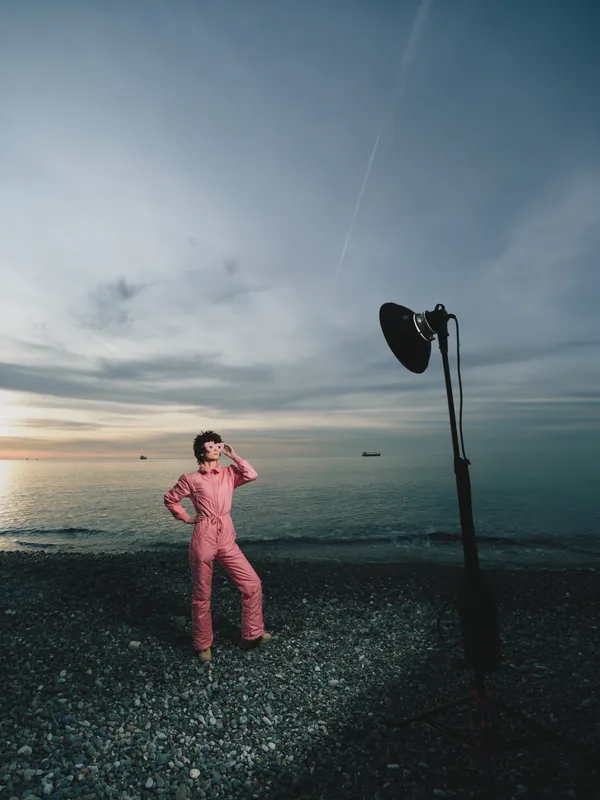 Model in a pink jumpsuit poses on a pebbled beach with a tall studio light against a sea sunset, blending fashion outdoor photography and dramatic coastline mood