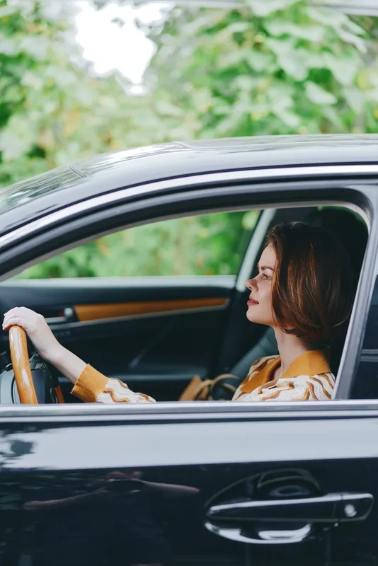 A woman sits in a sleek car, hands on the steering wheel, side profile visible through the window.