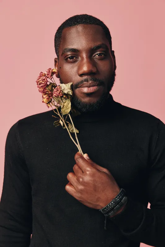 African man with dark skin holding dried flowers near face in creative conceptual visual style. Portrait with black sweater on pink background for artistic expression and emotion.