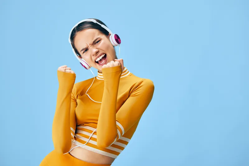 Energetic young woman in a yellow athletic crop top with long sleeves and headphones celebrates a fitness victory, pumping her fists with joyful expression against a clean pastel blue studio