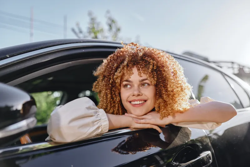 smiling woman with curly hair leans out of car window on sunny day, enjoying fresh air and bright outdoors, casual white blouse, natural joy expression