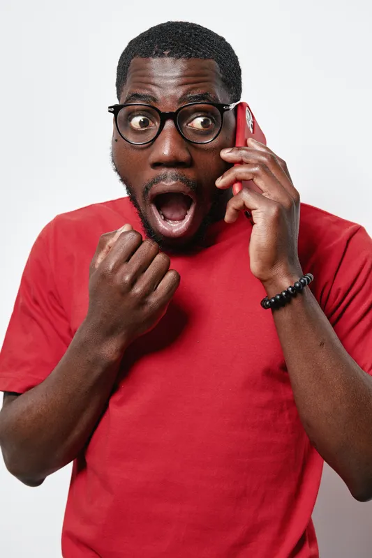 Black man with dark skin and glasses wearing red shirt talking on phone with shocked expression and open mouth isolated on white background. Surprised African American male in casual wear.