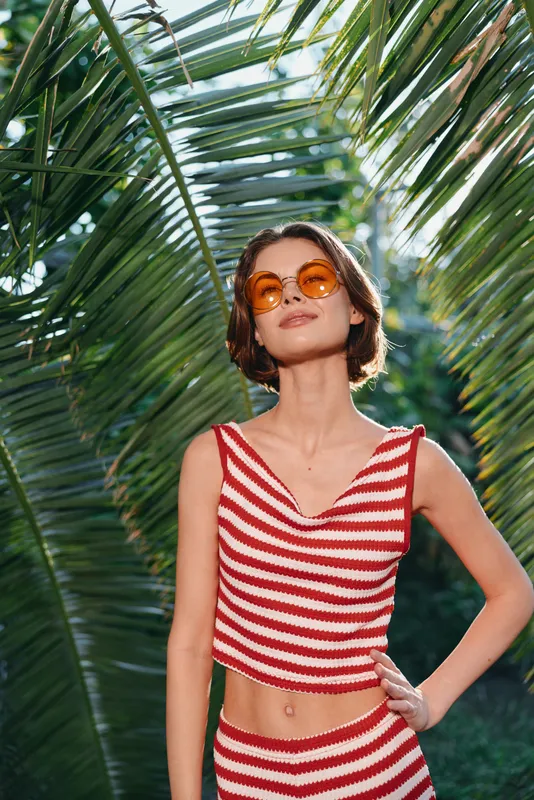 A stylish young woman wearing a red and white striped crop top and matching bottoms stands outdoors beneath lush palm leaves. She wears round sunglasses and poses confidently.