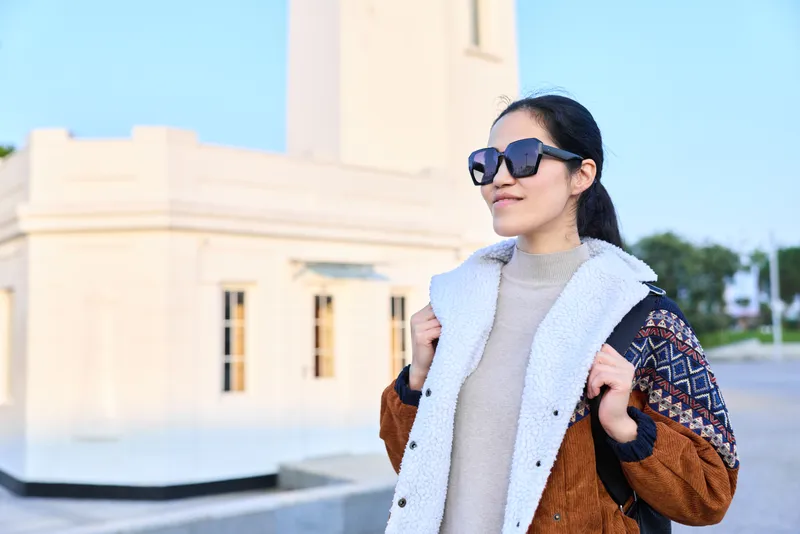 Woman wearing sunglasses, layered coat and fluffy scarf stands outdoors near a light architectural building, stylish and confident, capturing a fashion moment in bright daytime light.