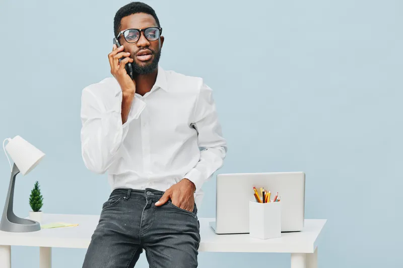Professional young African American man wearing white shirt and glasses, standing near a desk, talking on mobile phone, in a modern office environment with minimalist design, workspace, lifestyle conc