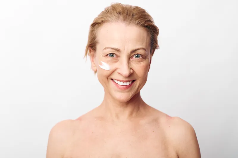 Smiling middle aged woman with cream on face looks at camera, glowing healthy skin and natural beauty. Concept for skincare, wellness, anti aging and self care routine.
