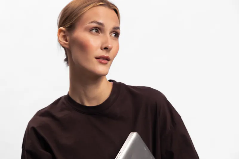 Young woman with light hair wearing dark shirt holds laptop, looking sideways. Studio portrait on white background. Concept of technology, work, and casual style.