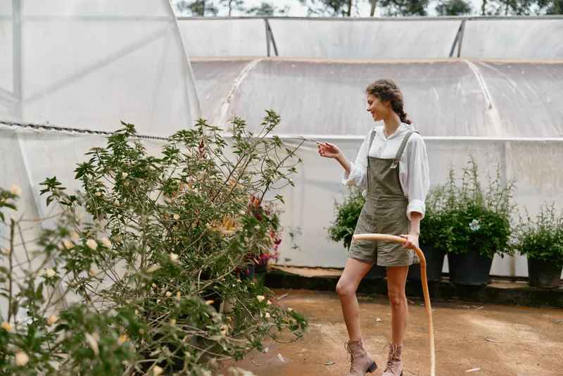 Young woman in green overalls watering plants in a greenhouse with a joyful expression, showcasing a sustainable gardening lifestyle surrounded by vibrant greenery
