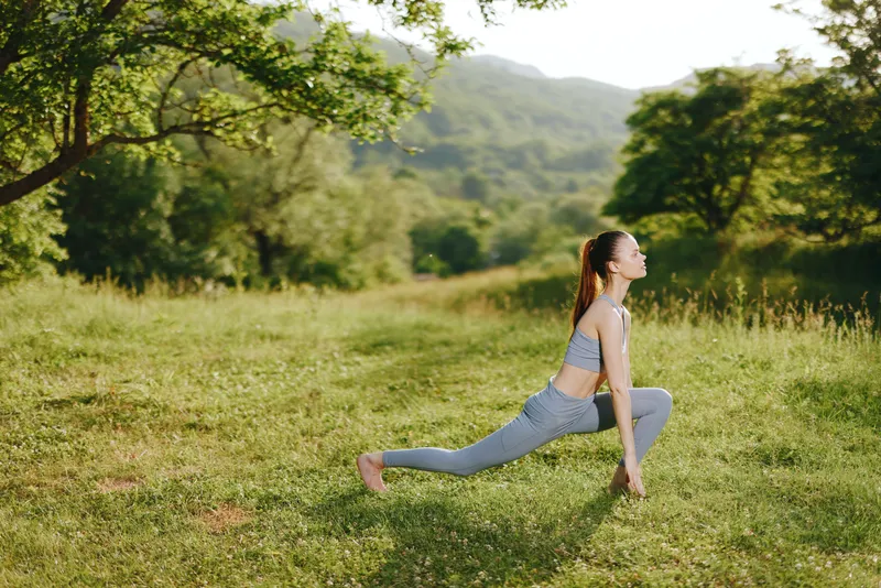 Young woman practicing yoga in nature, wearing gray activewear, conveying serenity and focus in a lush green landscape, promoting healthy lifestyle and wellness