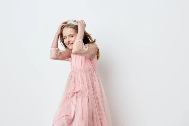 Smiling girl in a pink dress with a playful pose against a minimalist white background, showcasing joy and youthfulness in a studio setting