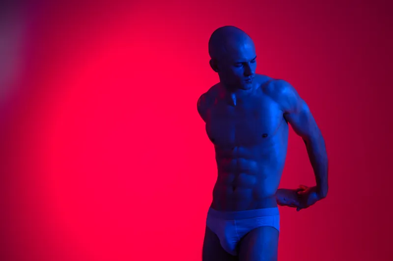 Athletic man posing under colorful lights, showcasing strong physique and confident posture in a striking visual display. The image features a vibrant color palette.