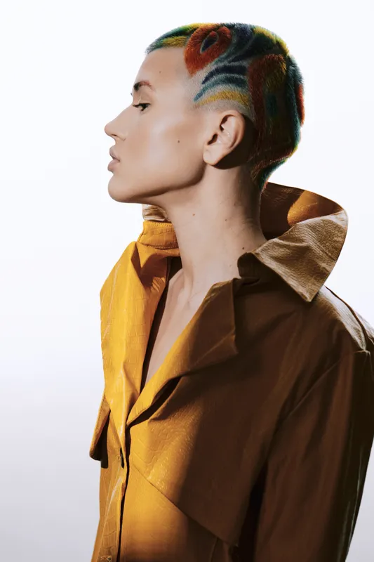 Woman with colorful shaved hair and yellow coat, showcasing modern artistic style, clean background, and unique fashion expression in profile portrait.