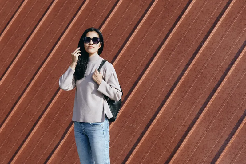 Woman with sunglasses and backpack standing against diagonal wooden panels in casual urban fashion, showcasing street style and modern confidence, with relaxed pose and soft light.