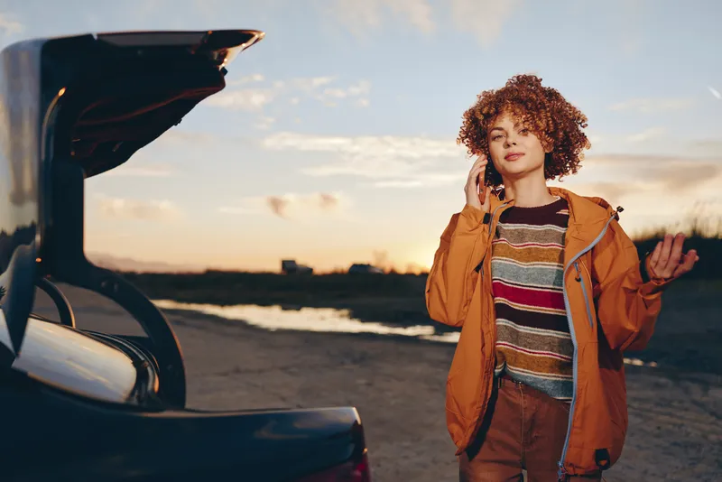 Woman lifestyle standing by open car trunk near beach at sunset, curly hair, casual jacket and backpack, relaxed travel mood, wind in hair, golden sky, coastal escape