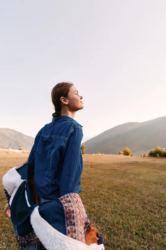 Woman in denim jacket stands in open field near mountains at sunset, enjoying nature and relaxation outdoors in a contemplative portrait with soft light and peaceful mood.