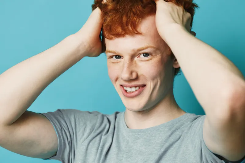 Young redhead man with freckles wearing gray t shirt smiling and holding hands on head acting playful and happy with bright expression on blue background.