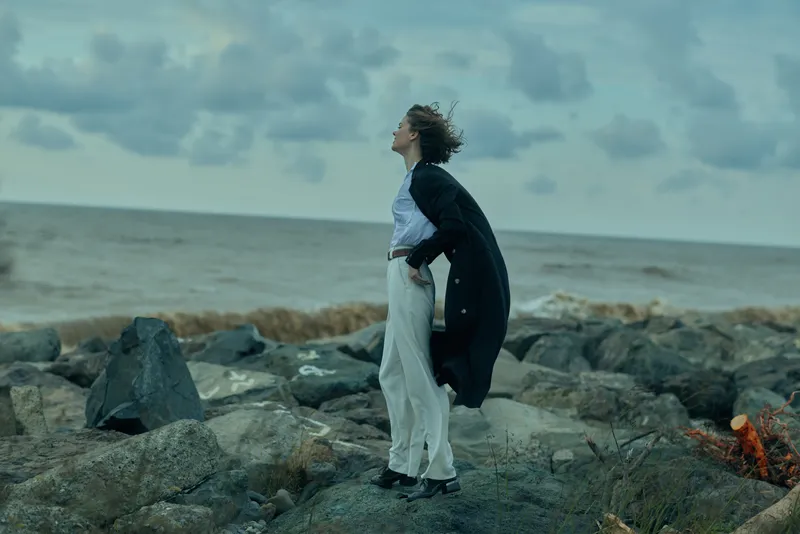 Windblown cape, thoughtful woman on a rocky shore by the sea, flowing coat and light trousers, moody sky, coastal fashion mood, travel and nature photography