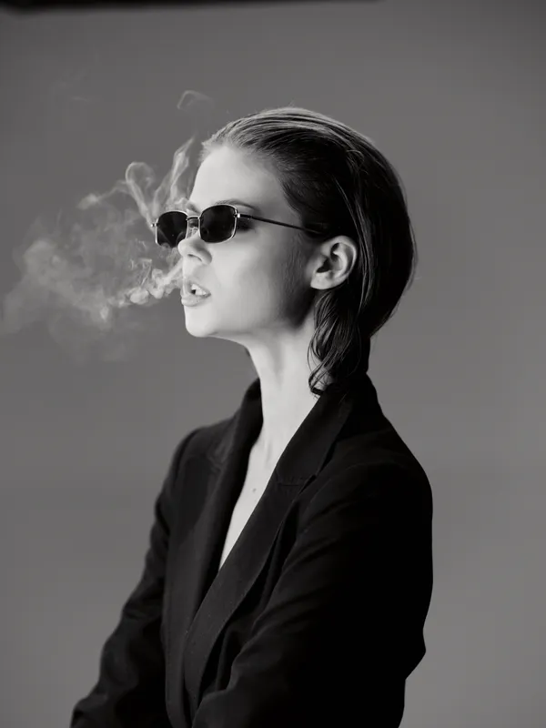 Black and white portrait of a stylish woman in sunglasses exhaling smoke, wearing a dark blazer with a confident expression.