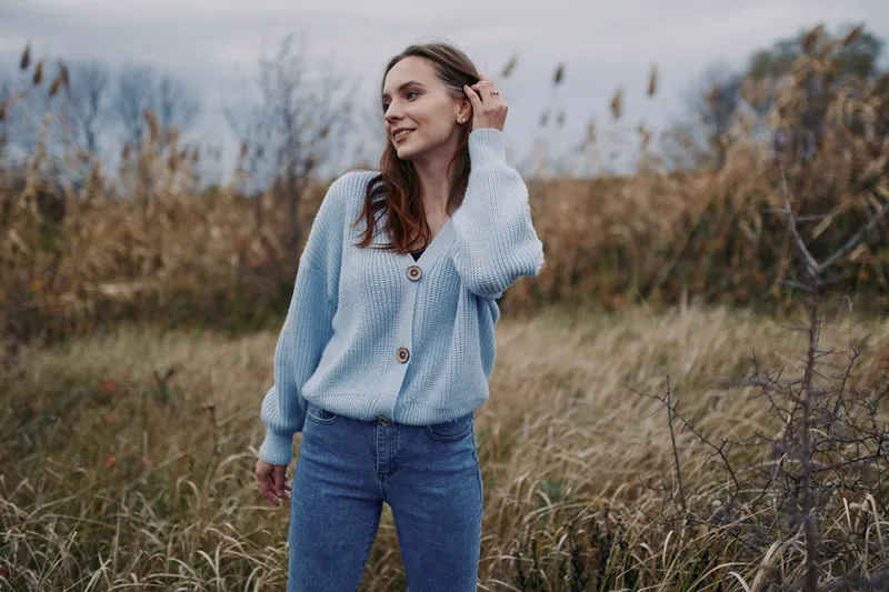 A candid portrait of a woman in a soft blue knitted cardigan standing in a natural field, conveying authenticity and confident presence in gentle light.