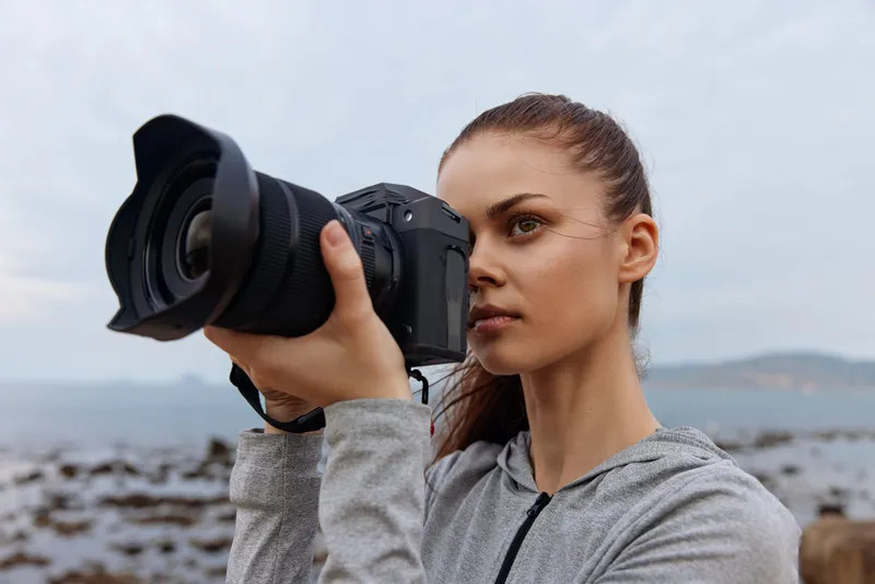 Young woman with a camera capturing a scenic landscape, showcasing her passion for photography with a focused expression at the beach