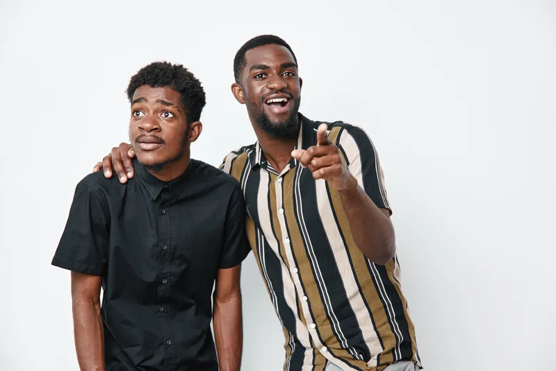 Two African American men with dark skin standing together on white background one pointing and smiling while the other looks surprised and thoughtful.