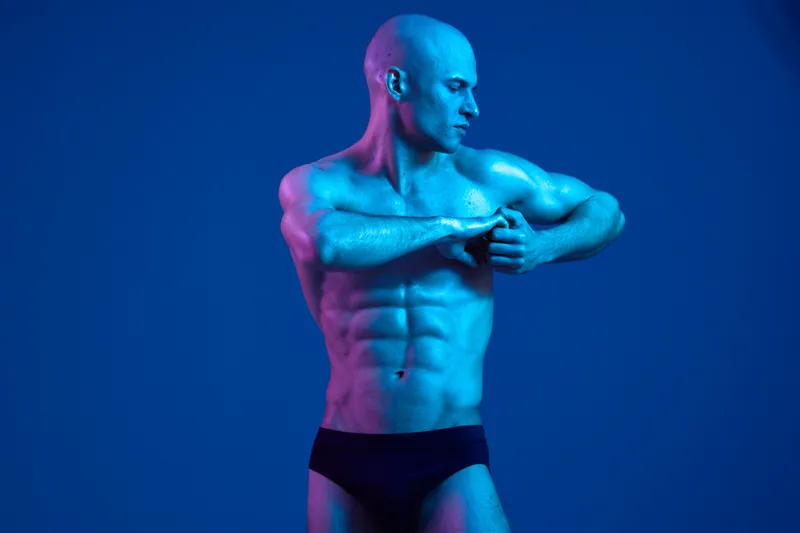 Athletic man posing in a vibrant blue light, showcasing his muscular physique and confidence. The image emphasizes strength and determination in a dynamic way.