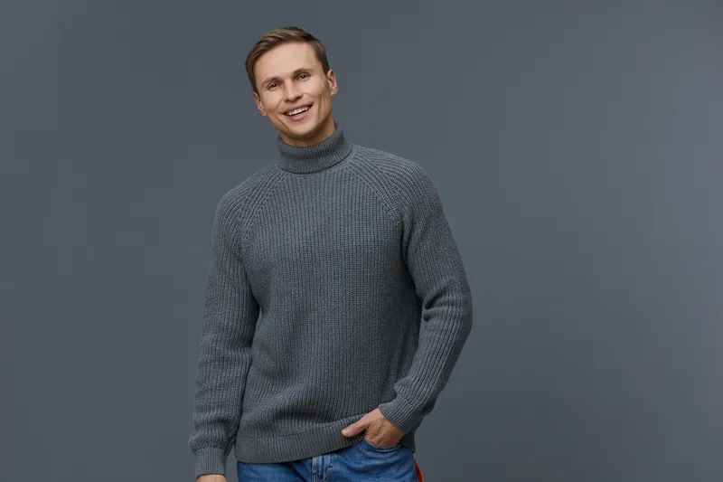 Smiling man in casual gray sweater standing against plain gray background, looking at camera with friendly expression, relaxed posture, modern fashion style, studio portrait, people lifestyle concept.