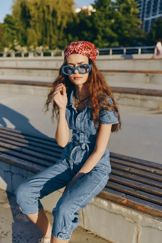 Lifestyle woman in modern boho-western denim street style wearing sunglasses and red bandana, captured with film color tones, sitting outdoors in natural light.