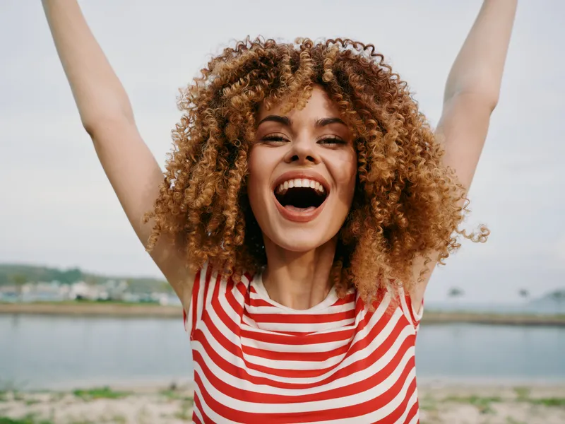 Joyful young woman with curly hair raises arms in celebration, wearing a red striped shirt at a sunny beach. A candid portrait full of life, warmth, and energy.