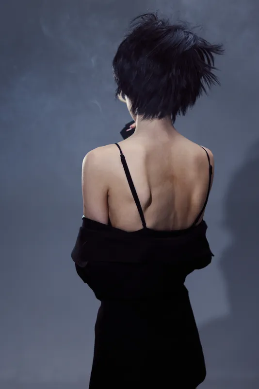 Woman with short hair backside portrait in dark dress, moody lighting, and minimalist background.