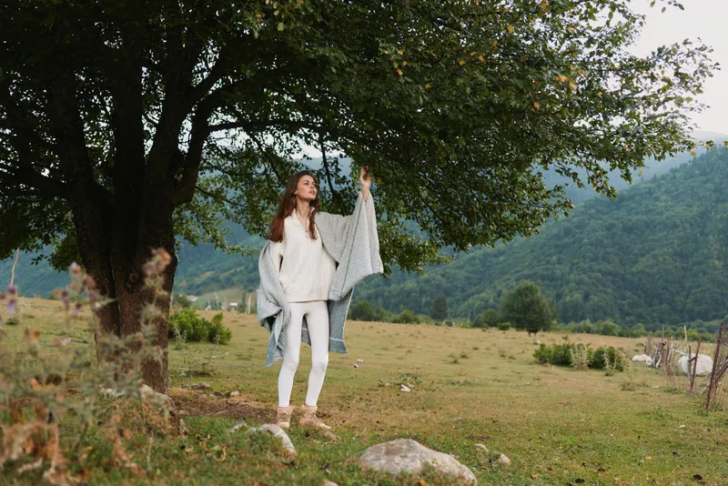 Woman in white casual clothing stands under a large tree in a rural landscape, enjoying fresh air and sunshine. A peaceful lifestyle moment captured in natural daylight.