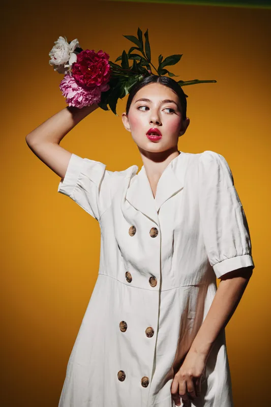 Young female fashion model with floral headpiece poses against a yellow background wearing a white buttoned dress. Studio beauty shot with creative styling concept and vibrant colors.